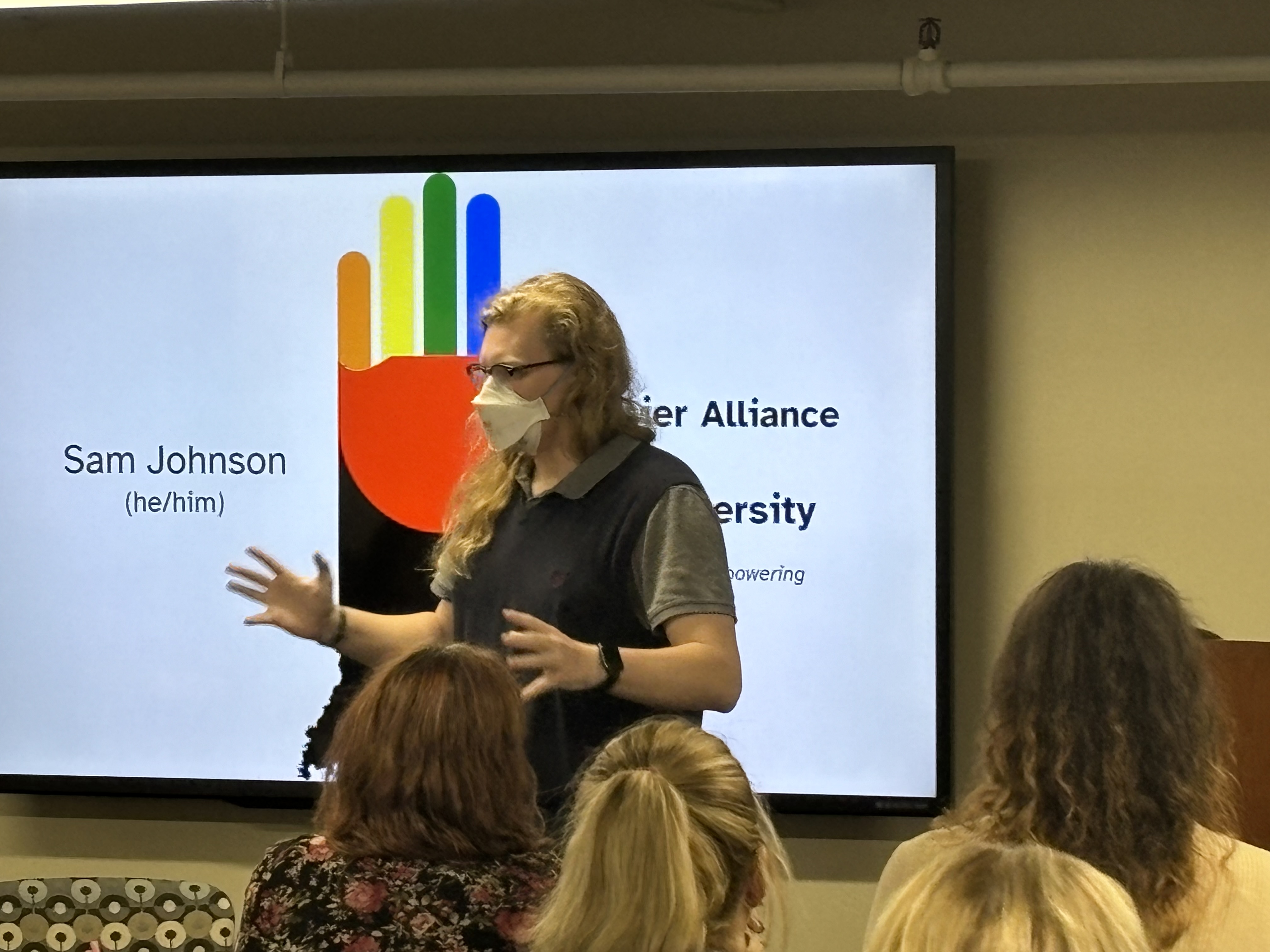 Standing and talking before a seated audience is, Sam Johnson, a white person with long blonde hair, wearing a glasses and respirator on his face with a grey polo shirt and a navy blue sweater vest. Behind him, a presentation slide displays the HAND logo (a combination of a rainbow-colored hand and the outline of Indiana) and his name and pronouns (he/him).